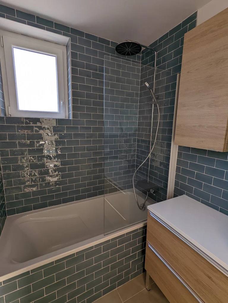 Salle de bain à Rezé après rénovation — baignoire avec pare-bain, douche encastrée, faïence métro bleu canard et meuble haut bois clair